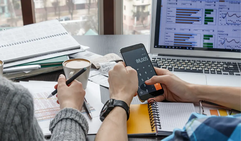 PPC_PAID (Pay-per-click-annonsering) - Profit Media_1024x600 Bild på två persone bakifrån. De sitter vid ett skrivbord med en mobil och en dator med massa papper och staitsik på datorn. Beräkning av annonser sker på mobilen.