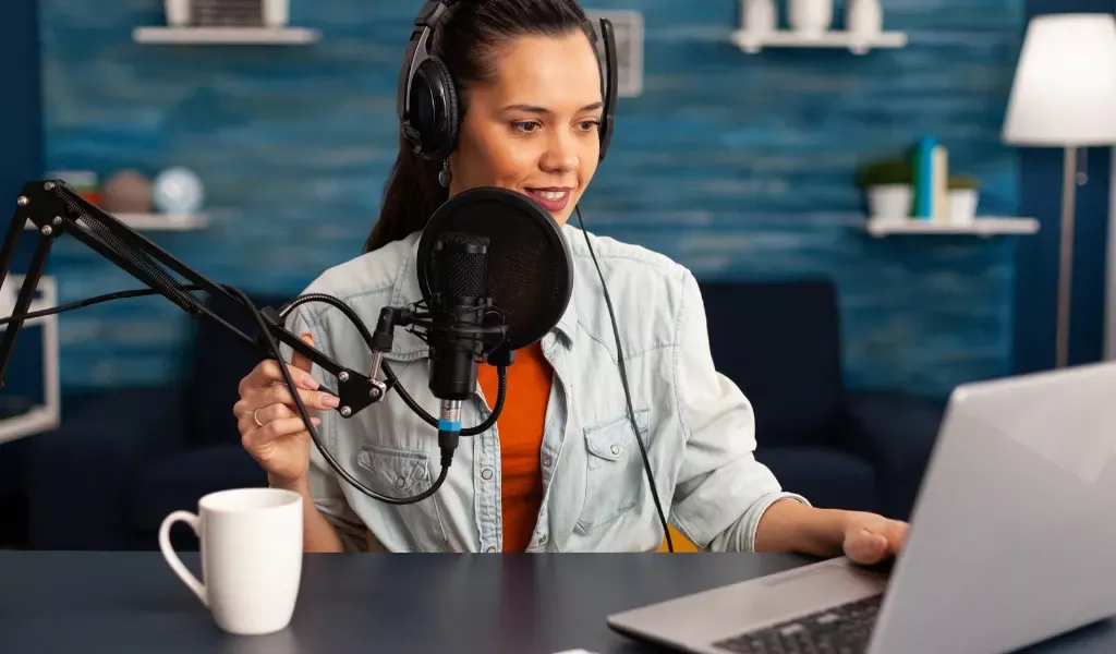 Bild på kvinna som sitter vid datorn med ett headset och spelar in en video via mikrofon och dator. På bordet har hon en kaffekopp och en laptop som hon tittar på.